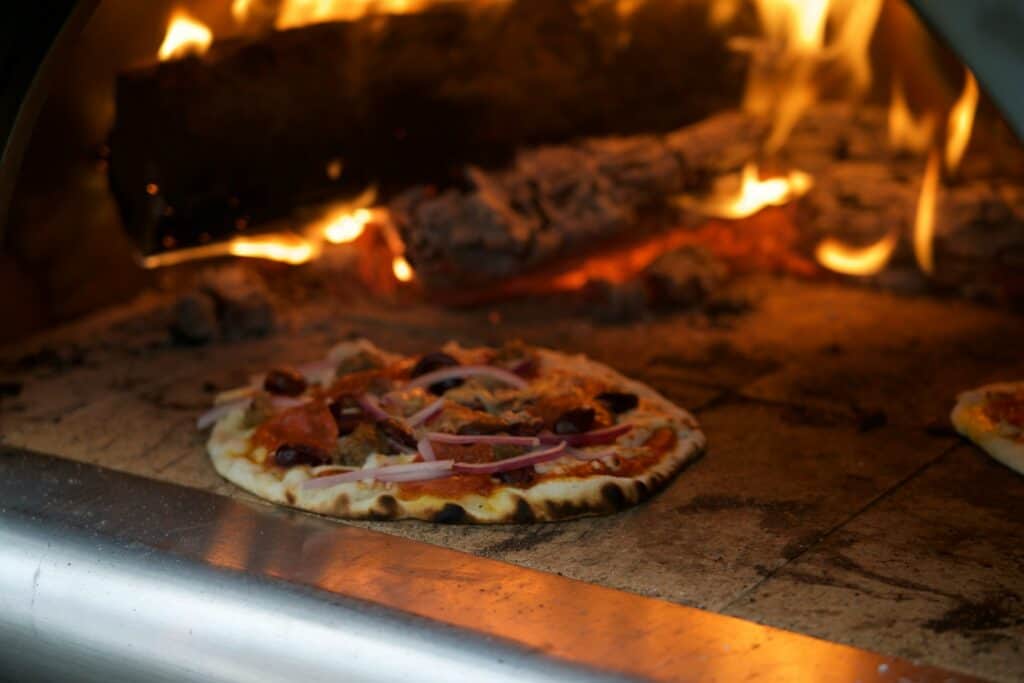 Wood-fired pizza cooking inside a stone oven, with open flames and glowing embers.