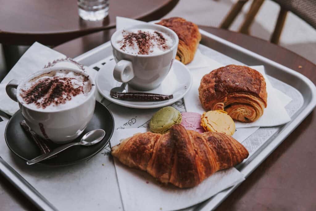 Two cappuccinos and assorted pastries including croissants and macarons on a café tray — breakfast at a local Douglas coffee shop.